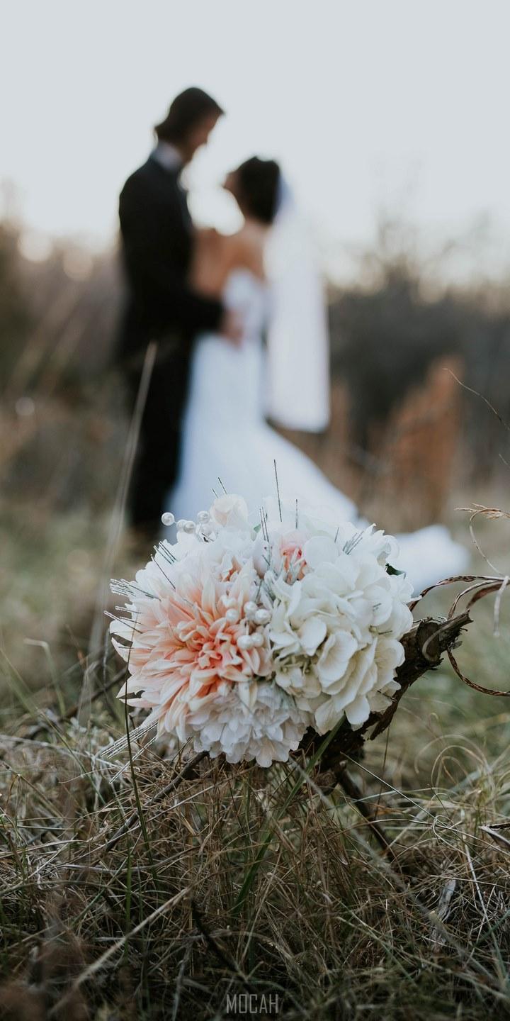 HD wallpaper, Cat S52 Wallpaper Hd, Wedding Evening, 720X1440, A Bouquet Sits In Tall Grass While A Married Couple Embraces In The Fuzzy Background